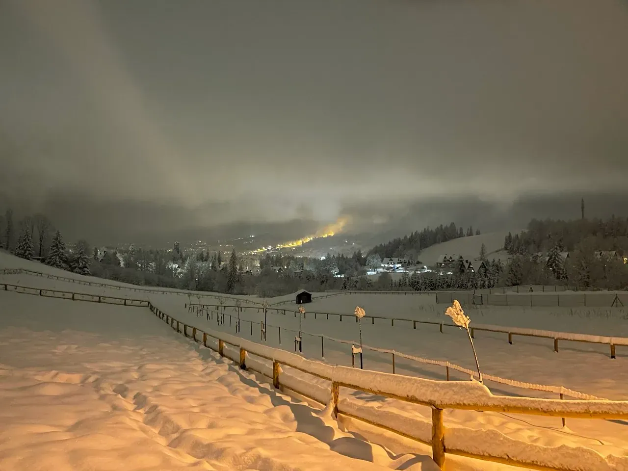 Snowy meadow and wooden fence on a winter evening, with the lights of Zakopane under low cloud